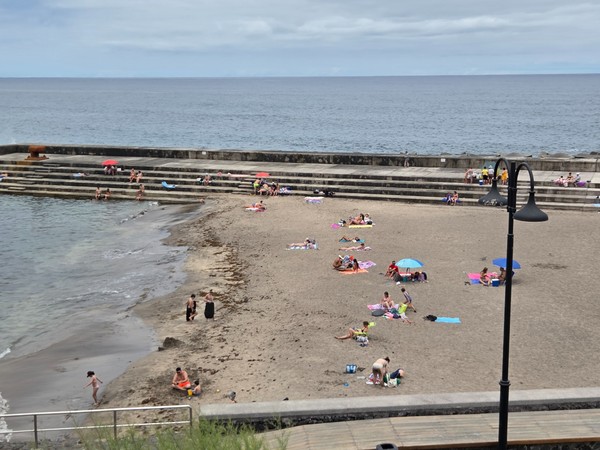 Plage de Bajamar avec baigneurs, gradins en béton et mer calme par ciel nuageux.