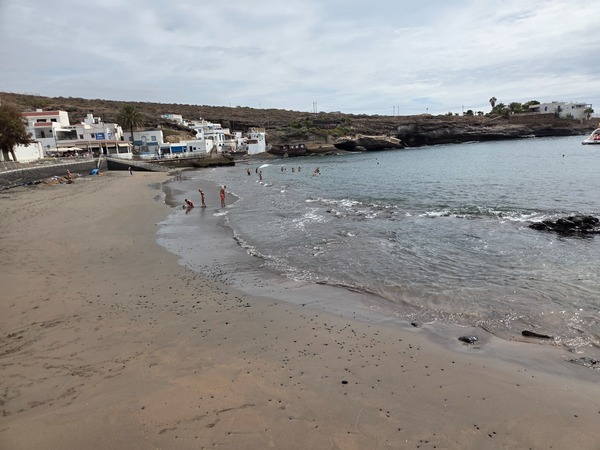 Plage de sable sombre d’El Puertito avec baigneurs et bord de mer tranquille.