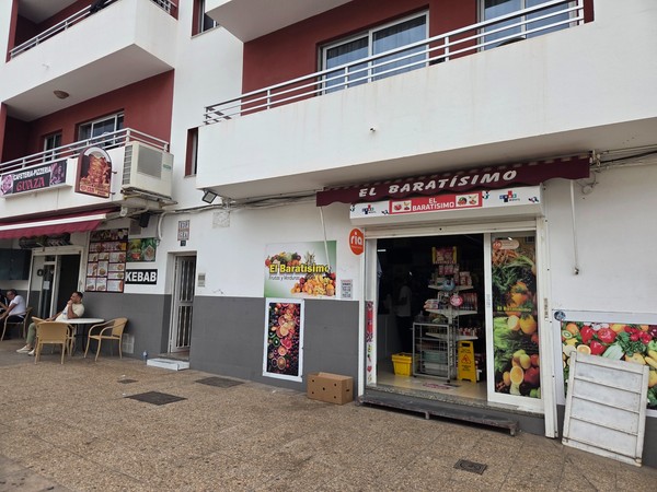 Entrée du magasin El Baratísimo à Guaza avec des affiches de fruits et légumes, un service de transfert d’argent et un snack kebab voisin avec terrasse.