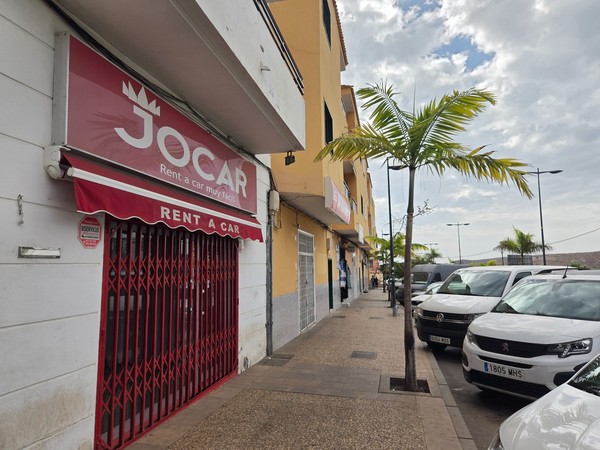 Agence de location de voitures Jocar à Guaza avec une enseigne rouge, des palmiers et une rangée de véhicules stationnés devant le bâtiment.