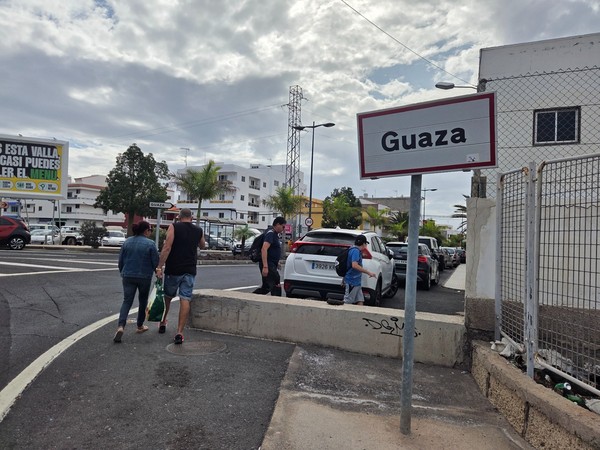 Panneau routier indiquant l’entrée de Guaza à Tenerife, près d’une route fréquentée avec des voitures garées et des passants.
