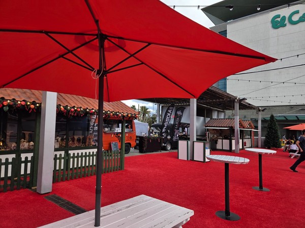 Espace détente du marché de Noël El Corte Inglés avec grandes toiles rouges, tables hautes et camionnette de restauration.