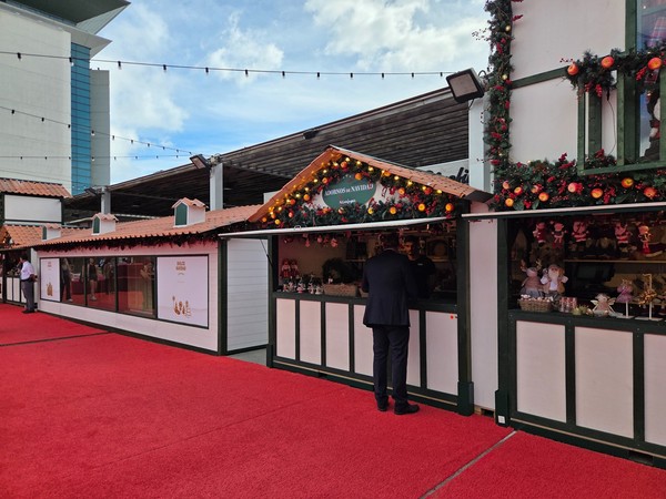 Alignement de stands de cadeaux de Noël décorés de lumières et de guirlandes devant El Corte Inglés, avec des visiteurs en train de faire leurs achats.