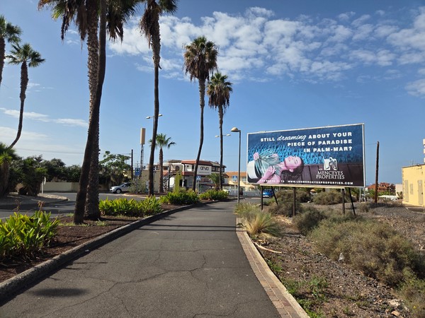 Grand panneau publicitaire d’une agence immobilière Los Menceyes Properties annonçant des propriétés à Palm-Mar le long d’un chemin piétonnier avec palmiers.