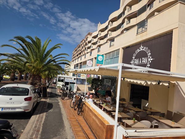 Terrasse ensoleillée du café-boulangerie Pan a Pan à Palm-Mar, avec des clients assis et une rangée de palmiers le long de la rue.