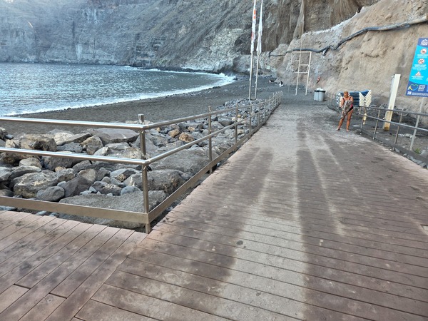 Promenade en bois avec garde-corps menant directement au sable noir de la plage.