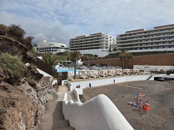 Vue depuis les escaliers sur Playa El Pinque : lits balinais de l’hôtel, piscine turquoise, palmiers et appartements en terrasse.