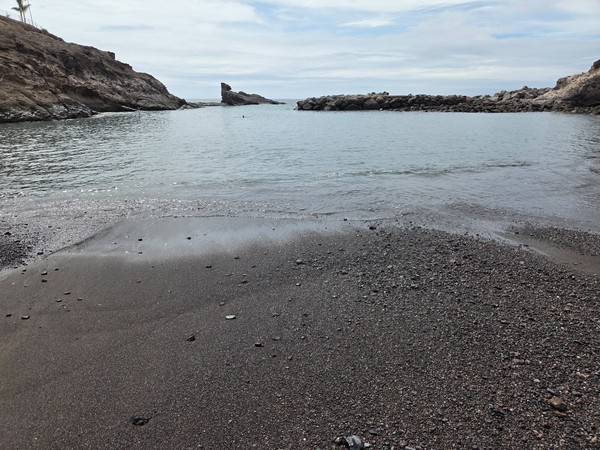 Gros plan sur le sable noir et les galets de Playa El Pinque avec l’eau calme de la crique au second plan.