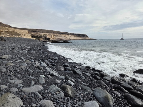 Vue depuis la grève de Playa Pardela vers l’horizon, vagues blanches sur galets noirs et un voilier au large.