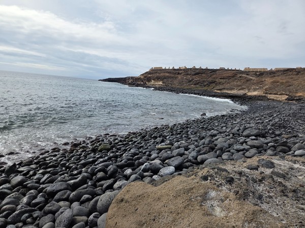 Panorama de Playa Pardela montrant la courbe de la baie couverte de galets noirs et les falaises claires au fond.