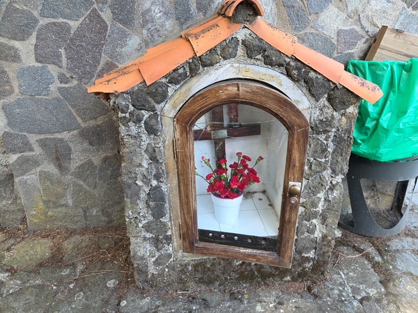 Petit oratoire en pierre avec une croix en bois et un pot de fleurs rouges derrière une vitre.